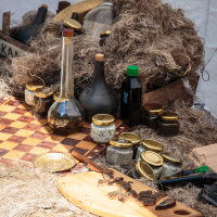 A rustic display on a wooden table at Kalnciema kvartāls featuring various items. There are jars with golden lids, dark-colored bottles, a wooden board with slices of dried meat or bread, and several old-fashioned, cork-topped glass bottles. The straw-covered table creates a vintage, artisanal atmosphere.