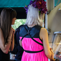 A woman with long, grey hair adorned with large, dark purple flowers stands at an outdoor market stall in Kalnciema kvartāls. She is wearing a bright pink dress with a black leather collar and belt. Another woman with long brown hair and sunglasses stands beside her, looking at the Modes Diena stall.
