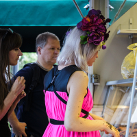 A woman with platinum blonde hair, adorned with a large flower headpiece, wears a vibrant pink dress and black backpack. She stands inside a shop at Kalnciema kvartāla during modes diena, intently examining items in a display case. Behind her are a man with a black shirt and a woman with long, dark hair wearing glasses.