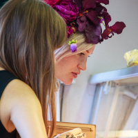 Two women are closely looking into a display case at Kalnciema kvartāla. One has long brown hair, while the other has blonde hair adorned with a large flower headpiece in shades of purple and red. The brown-haired woman has an intricate black henna design on her left hand and is holding a beige purse.