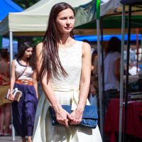 A woman with long dark hair walks through the outdoor market at Kalnciema kvartāla. She wears a sleeveless white dress and holds a blue clutch. She is in focus, while other people and market stalls with colorful canopies are visible in the background. The weather appears to be sunny on this modes diena.