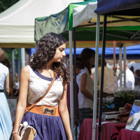 A woman with long curly hair, wearing a polka dot sleeveless top and a navy blue skirt with a wide belt, holds a brown paper bag in each hand as she looks to her left. She stands in front of vendor stalls at Kalnciema kvartāla modes diena, with canopy tents providing shade and people browsing in the background.