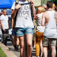A young girl walks outdoors, carrying a backpack. She wears a white t-shirt with a print, denim shorts, and blue sneakers. Sunglasses rest on her head. Behind her, people gather at market stalls with colorful tents in blue, green, and beige hues of Kalnciema kvartāla on a lively diena.