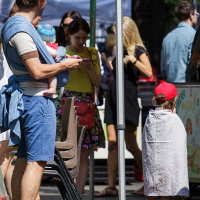 Adults and children enjoy the festivities at a Kalnciema kvartāla event. A man in a blue shirt, blue shorts, and sandals holds a baby in a floral hat and blue wrap. A boy in a red cap and white cape stands near a sign as people in colorful clothing gather under blue and yellow tents in the background.