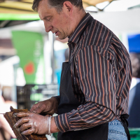 A man, wearing a black apron over a striped shirt and jeans, is working at Kalnciema kvartāls market. He is handling a rack of ribs while wearing plastic gloves. The background features market stalls with green and blue canopies and blurred patrons on modes diena.