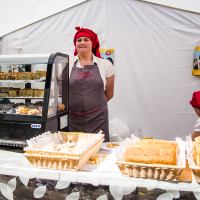 Two women are in a white tent bakery stand at the Kalnciema kvartāla market. One woman stands behind a display case filled with various pastries while wearing a red headscarf and gray apron. The other woman, also in a red headscarf, attends to something on the side. Baskets of bread and pastries are on the table in front.