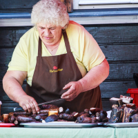 A woman with curly blonde hair, wearing a yellow shirt and a brown apron, prepares assortments of sausages and meat at Kalnciema kvartāla's wooden counter during modes diena. Various meats and containers are spread out in front of her. The background showcases a wooden wall and a red plastic container by her side.