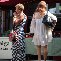 Two women stand near a market stall at Kalnciema kvartāla. The woman on the left has short hair, wears a black and white striped maxi dress, and holds a floral bag. The woman on the right has long hair, wears a white off-the-shoulder dress, carries a black bag, and wears heels. The market stalls have red canopies.