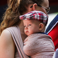 A woman with brown hair in a ponytail, wearing glasses, carries a baby in a patterned wrap at modes diena in Kalnciema kvartāla. The baby, wearing a red and white plaid cap, looks to the side with a curious expression. The background is blurred with hints of red and blue.