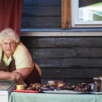 A person with curly blonde hair wearing a light yellow shirt and brown apron leans on a counter at an outdoor market stall during Kalnciema kvartāla modes diena. Various smoked meats are displayed on the counter, and a red curtain and dark wooden wall with a window are in the background.