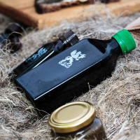 A black rectangular bottle with a green cap and a white emblem lies on a bed of straw next to a smaller amber bottle with a brown cap. In the foreground, a small jar with a golden lid is visible. The scene evokes the rustic charm of Kalnciema kvartāls during modes diena, with bread slices spread on a wooden board in the background.