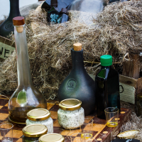 A rustic display featuring various bottles and jars on a wooden chessboard at Kalnciema kvartāla captures the essence of modes diena. Tall, dark glass bottles with green caps and different shapes are surrounded by straw. Glass jars with golden lids contain various substances. A small, open notebook and two short glasses with a light yellow liquid are at the front.