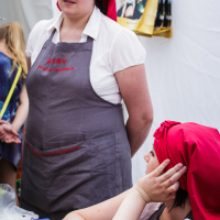 Two women, wearing red headscarves and gray aprons, are seen in a booth at Kalnciema kvartāla. The standing woman looks ahead while the seated woman rests her head on her hand, gazing to the side. A white backdrop frames them with various items on the table and a bag hanging in the background, capturing a moment of modes diena.