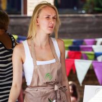 A woman with long blonde hair, wearing a white shirt and a beige apron with a green logo, stands behind a catering stall at the Kalnciema modes diena. The background shows colorful triangular buntings and another person in a striped shirt. A foil food tray, cup, and menu rest on the counter.
