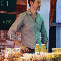 A man stands behind a market stall with various packaged goods and two bottles of liquid on display in Kalnciema kvartāla. He wears a light blue shirt and has sunglasses perched on his head. The stall is under a green canopy, with signs in the background, and some packs appear to contain dried fruits.