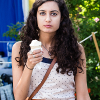 A woman with long, dark, curly hair stands outdoors at Kalnciema kvartāls, holding an ice cream cone with her right hand. She is wearing a white polka-dotted sleeveless top, a brown belt with a bow buckle, and a crossbody bag. The background includes greenery and a partially visible tent with a blue and yellow covering, suggesting she's enjoying modes diena.