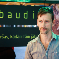 A man with short brown hair and sunglasses resting on his head stands in front of a tented booth at Kalnciema kvartāls. The sign behind him reads "baudi" and "Garšas, kādām tām jābūt." He is wearing a blue button-up shirt and appears to be looking slightly to the side, enjoying the modes diena atmosphere.