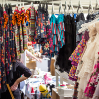 A colorful market stall at Kalnciema kvartāla displays an array of children's clothing. On the left side, rows of bright floral and striped dresses hang neatly, while the right side shows pastel-toned, lacy dresses. In the background, shoppers peruse more items under a white canopy on this lively modes diena.