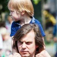 A man with long brown hair carries a small child with blond hair on his shoulders at Kalnciema kvartāla. The child, wearing a navy blue shirt, looks off to the side while the man, appearing focused, grips the child's legs for support. The blurred background shows people and greenery.