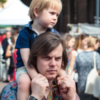 A man with medium-length hair, dressed in a colorful, patterned shirt, carries a small blonde child on his shoulders. The child is wearing a blue shirt and looking intently into the distance. In the blurred background of Kalnciema kvartāls, a crowd and some trees suggest it's modes diena in this public outdoor setting.