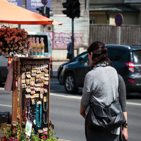 A person with dark hair and a gray top is standing in front of an outdoor market stall adorned with various jewelry items such as necklaces and earrings. The stall, part of Kalnciema kvartāla's lively scene, is shaded by an orange umbrella, with flowers at its base. A street with cars and traffic signals is in the background on this modes diena.
