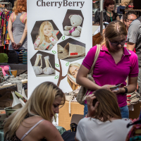 A bustling market scene at Kalnciema kvartāls with a prominent banner displaying "Made in Latvia, CherryBerry" featuring images of children’s toys. Shoppers, including a woman in a pink shirt and another in a white top, browse various stalls filled with handmade items and crafts on this modes diena.