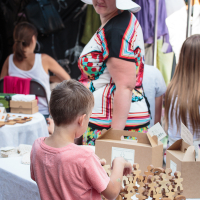 A young boy in a pink shirt examines wooden puzzle pieces at a market stall in Kalnciema kvartāla. A woman in a colorful dress and white hat with floral patterns is smiling in the background. Other shoppers and stalls with various items can be seen, contributing to a busy, vibrant atmosphere on this modes diena.