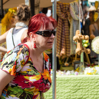 A woman with red hair, wearing sunglasses, a bright floral shirt, and red earrings is browsing items at the Kalnciema kvartāla market during the modes diena event. Behind her are stalls with colorful fabric and various goods on display. The background also shows other shoppers and a yellow canopy.