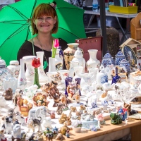 A smiling woman with red hair sits under a green umbrella at Kalnciema kvartāla market stall displaying various ceramic and porcelain items, including teapots, vases, figurines, and animal sculptures. On this vibrant modes diena, another person is partially visible to the right amidst more displayed items.