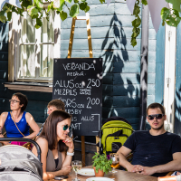 People are sitting at an outdoor cafe with a rustic wooden exterior and leafy green plants overhead, enjoying the afternoon at Kalnciema kvartāls. A chalkboard menu details drink options, listing beer and craft ales. One woman is drinking from a glass, while others are talking and relaxing around a wooden table in the sunlit area.