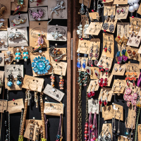A display of various colorful earrings and necklaces mounted on cardboard tags, hanging on a dark wooden rack at Kalnciema kvartāla. The jewelry, marked "Pure Metal," features a mix of floral patterns, animal shapes, and geometric styles. This dense arrangement is truly visually striking and embodies the essence of Modes Diena.