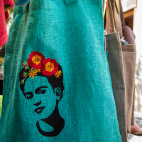 A turquoise fabric tote bag displays a black artistic print of a person's face, adorned with colorful flowers in their hair. The bag hangs amongst other items at the Kalnciema outdoor market, with glimpses of greenery and wooden elements in the background.