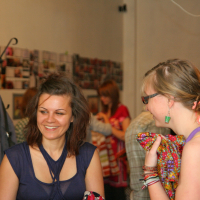 Two women are engaged in talka in a warmly lit indoor setting. The woman on the left, smiling, has shoulder-length brown hair and wears a sleeveless navy blue top. The woman on the right has blonde hair, wears glasses, and holds colorful fabric. They are among other people and some wall decor.