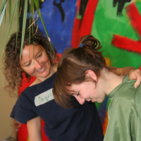 A person with curly hair and a shirt that has a name tag is smiling and leaning toward another person with brown hair, who is laughing and looking down. They are standing in front of a colorful abstract mural, immersed in their modes of communication. Some green leaves hang down from the top of the image.