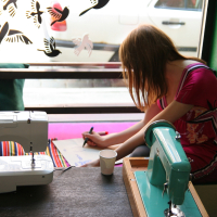 A person wearing a pink dress with a purple and white pattern is sitting on a purple cushioned bench by a window, engaging in modes talka. They are writing on a piece of paper on their lap. In front of them are white and teal sewing machines on a black table, while bird decals adorn the window.