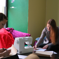 Two women focus on their work inside a room with green and yellow walls. One woman writes notes at a desk with a sewing machine and fabric, while the other, seated nearby with a notebook, writes intently. They sit on a couch and chair, respectively, near a window letting in natural light—modes talka filling the air.