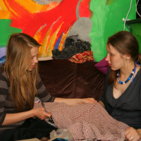 Two women are sitting on a couch in front of a colorful abstract mural, engaging in modes talka as they examine a piece of checkered fabric. One woman has long, straight blonde hair and wears a striped dress. The other woman, with brown hair pulled back, dons a dark gray dress with a blue beaded necklace.