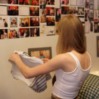 A person with long blonde hair, wearing a white tank top and beige pants, stands facing a wall. They hold a striped piece of clothing in both hands. The wall, adorned with a collage of various photographs and framed images, exudes Modes talka vibes. The room appears cozy with muted lighting.