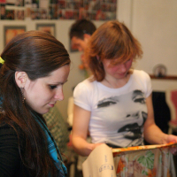 Two women are engaged in an activity indoors. The woman on the left, with long dark hair and a turquoise scarf, seems to be in deep modes talka with her friend. The woman on the right, wearing a face-printed white T-shirt and holding a beige patterned cloth, listens intently. Blurred images adorn the background walls.
