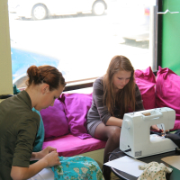 Two women are engaged in sewing activities in a bright room with large windows. One woman on the left hand-sews fabric adorned in blue patterns, while the woman on the right uses a white Modes sewing machine. The cozy room features pink and purple cushions, with paper cranes hanging in the window.