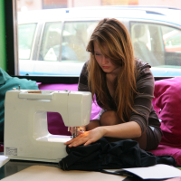 A woman with long hair is using a Modes talka sewing machine while seated on a magenta cushioned bench next to a green wall. She is focused on stitching fabric. There are papers and more fabric around her. A large window behind her shows parked cars, providing natural light to the workspace.