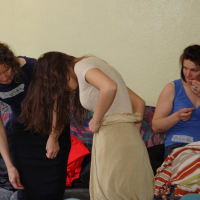 Three women are seated on a colorful couch. Two women on the left are bending over, examining something on the floor. The woman on the far left has curly hair and a blue top, and the middle woman has long hair and a beige skirt. The third woman on the right is sitting upright in a blue top, seemingly lost in talka modes of thought.
