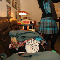 A woman in a plaid dress stands in a room arranging items, her face not visible. In front, a cluttered brown couch is piled high with various clothes. The room, decorated with hanging photos and art on the wall, features a red lamp beside other furniture and offers hints of retro modes.