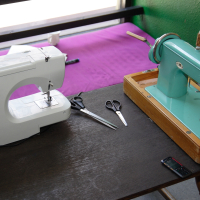 Two sewing machines on a wooden table: a modern white electric machine on the left and a vintage teal manual machine on the right. Scissors, thread, and a black mobile phone are also on the table, perfect for modes talka enthusiasts. A purple cloth and window are in the background.