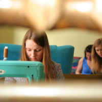 A young woman with long brown hair is focused on sewing with a turquoise machine in the foreground. Behind her, two other young women are engaged in talka on a couch. The scene is warmly lit, giving a cozy and intimate atmosphere, capturing their creative modes perfectly.