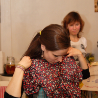 A woman with long brown hair, wearing a plaid shirt, holds up a red and blue patterned shirt in front of her, inspecting it closely. Another woman with short brown hair, wearing a light gray top, stands in the background observing. The setting appears to be indoors with various items around, creating a cozy Modes talka atmosphere.