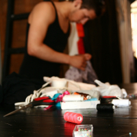 A person wearing a black tank top is standing and working with fabric in the background. The foreground is focused on a surface cluttered with sewing supplies, including a spool of pink thread, a container of pins, scissors, and various other sewing items—truly resembling the modes of a dedicated seamstress. A ladder leans against the wall.