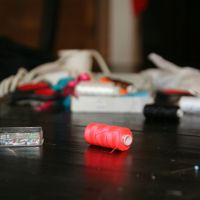 A close-up of a spool of red thread on a dark wooden table. In the background, the table is scattered with sewing supplies, including a box of multicolored pins, various spools of thread, fabric scraps, and an assortment of sewing notions—all slightly blurred to focus on the talka red thread spool.