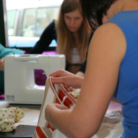 Two women are working with fabric and a sewing machine at a table. The woman in the foreground, with short dark hair and a blue sleeveless top, is holding fabric with red and white stripes. Meanwhile, the woman in the background, her long blonde hair cascading as she operates the sewing machine, participates in modes talka.