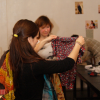 Two women hold and examine pieces of clothing in a room with light-colored walls and various pictures pinned to them. The foreground woman, sporting a colorful scarf, exudes Modes talka elegance. The background woman with shoulder-length brown hair holds up a patterned shirt.